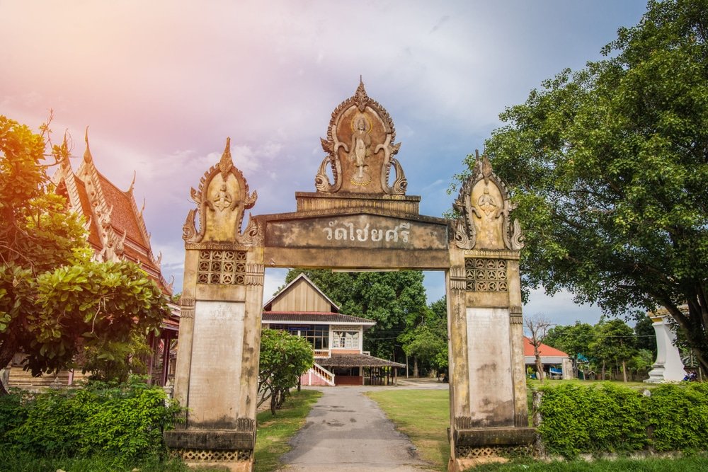 ประตูทางเข้าหินประดับประดาด้วยอักษรไทย นำไปสู่กลุ่มวัดแบบดั้งเดิม ซึ่งเป็นหนึ่งในสถานที่ท่องเที่ยวที่ต้องไปเยือนในขอนแก่น ประกอบด้วยอาคารหลังคาสีแดงและต้นไม้เขียวชอุ่มภายใต้ท้องฟ้าที่มีเมฆบางส่วน.