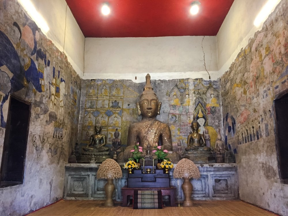 พระพุทธรูปขนาดใหญ่ตั้งอยู่ตรงกลาง โดยมีพระพุทธรูปขนาดเล็กและเครื่องบูชาอยู่ทั้งสองข้าง วางไว้บนผนังที่ประดับด้วยภาพจิตรกรรมฝาผนัง ภายในห้องพระที่มีเพดานสีแดงและพื้นไม้—สถานที่อันน่าประทับใจสำหรับผู้ที่ต้องการสำรวจสถานที่ท่องเที่ยวในขอนแก่น.