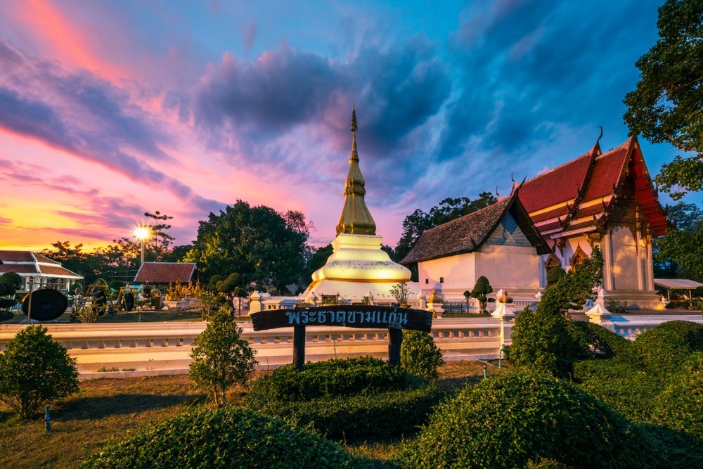 วัดพุทธที่มีพระธาตุทองคำและอาคารแบบดั้งเดิมในยามพระอาทิตย์ตกดิน พร้อมด้วยพุ่มไม้ที่ตัดแต่งอย่างสวยงาม ป้ายที่มีข้อความภาษาไทยอยู่ด้านหน้า—สถานที่ท่องเที่ยวที่เป็นสัญลักษณ์ของขอนแก่นสำหรับนักท่องเที่ยว.