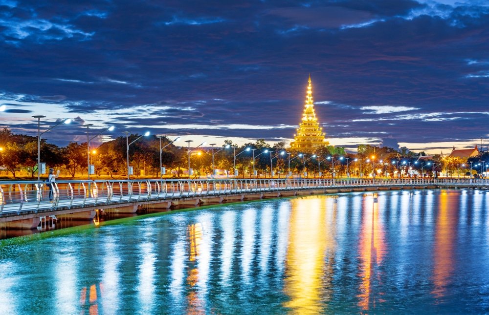 เจดีย์ทองคำที่ส่องแสงสว่างตั้งอยู่ริมแม่น้ำในยามพลบค่ำ แสงไฟสะท้อนบนผืนน้ำใต้ท้องฟ้าที่มีเมฆครึ้ม—เป็นหนึ่งในสถานที่ท่องเที่ยวที่ต้องไปเยือนในขอนแก่นสำหรับผู้ที่ต้องการสัมผัสประสบการณ์การท่องเที่ยวในขอนแก่น.