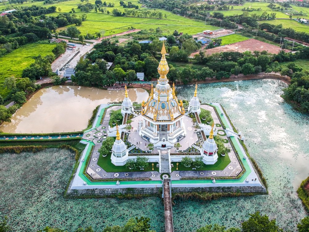 ภาพมุมสูงของวัดสีขาวและทองที่ล้อมรอบด้วยน้ำและทุ่งหญ้าเขียวขจี มีทางเดินนำไปสู่ทางเข้า—สถานที่ท่องเที่ยวที่ต้องไปเยือนในขอนแก่นสำหรับผู้ที่ต้องการสำรวจเที่ยวขอนแก่น.