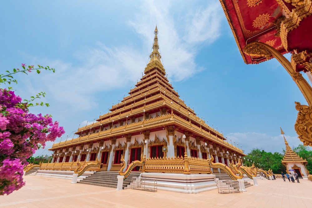 วัดพุทธขนาดใหญ่ที่ประดับประดาอย่างวิจิตรตระการตา มีรายละเอียดสีทองและหลังคาหลายชั้นตั้งอยู่ใต้ท้องฟ้าสีคราม ล้อมรอบด้วยดอกไม้และพืชพรรณเขียวขจี—สถานที่ท่องเที่ยวที่ไม่ควรพลาดในขอนแก่นสำหรับนักท่องเที่ยวที่มาสำรวจภูมิภาคนี้.