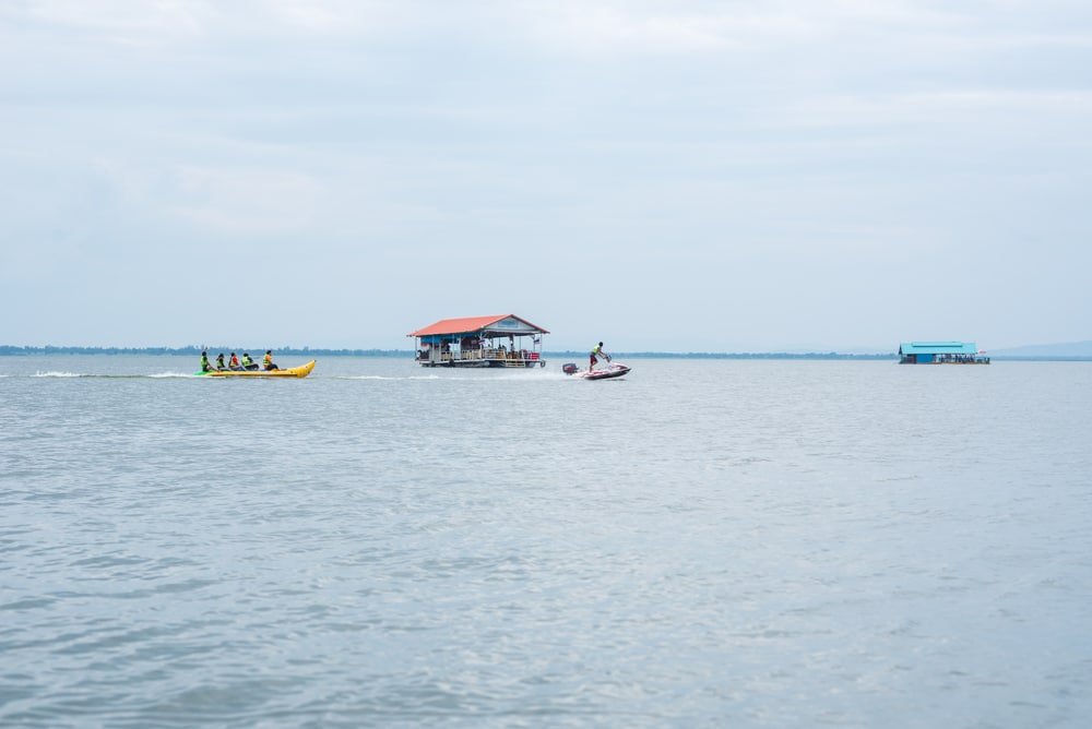 กลุ่มคนกำลังนั่งบนเรือกล้วยยางพองลมที่ถูกลากโดยเจ็ตสกีบนผืนน้ำขนาดใหญ่ โดยมีบ้านลอยน้ำสองหลังอยู่เบื้องหลังภายใต้ท้องฟ้าที่มีเมฆครึ้ม—กิจกรรมสุดตื่นเต้นที่สถานที่ท่องเที่ยวขอนแก่น.