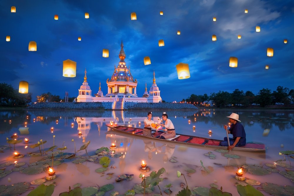 ผู้คนในเรือไม้ปล่อยโคมลอยขึ้นสู่ท้องฟ้าเหนือทะเลสาบใกล้กับวัดในยามพลบค่ำ โดยมีโคมไฟส่องแสงสว่างไสวบนท้องฟ้า—ภาพที่งดงามราวกับเวทมนตร์ซึ่งสะท้อนความงามของสถานที่ท่องเที่ยวในขอนแก่นและเสน่ห์ของการท่องเที่ยวในขอนแก่น.