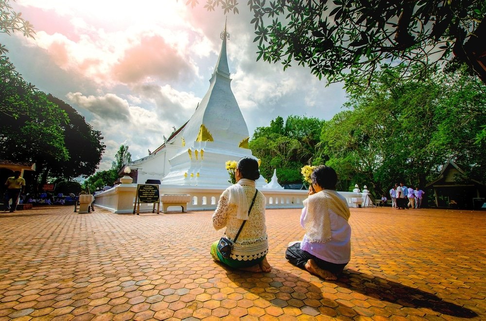 สองคนกำลังคุกเข่าและสวดมนต์อยู่หน้าเจดีย์สีขาวในวัด ที่ล้อมรอบด้วยต้นไม้ และใต้ท้องฟ้าที่มีเมฆบางส่วน—เป็นช่วงเวลาที่สงบสุข ซึ่งสะท้อนความงามของที่เที่ยวเชียงคาน  สำหรับใครก็ตามที่ต้องการมาสำรวจจุดหมายการท่องเที่ยวที่น่าทึ่งแห่งนี้.