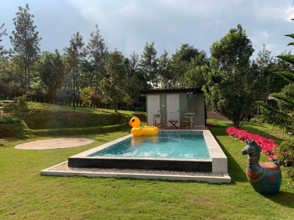 สระว่ายน้ำกลางแจ้งขนาดเล็กที่มีเป็ดยางสีเหลืองตัวใหญ่ ล้อมรอบด้วยหญ้า ต้นไม้ และดอกไม้สีสันสดใส มีเก้าอี้สองตัวและโต๊ะหนึ่งตัวตั้งอยู่ใกล้กับอาคารเล็กๆ ในพื้นหลัง — เป็นสถานที่ที่สมบูรณ์แบบสำหรับการพักผ่อนที่ รีสอร์ทเขาค้อ หลังจากได้สำรวจความงามของธรรมชาติในพื้นที่นี้แล้ว.