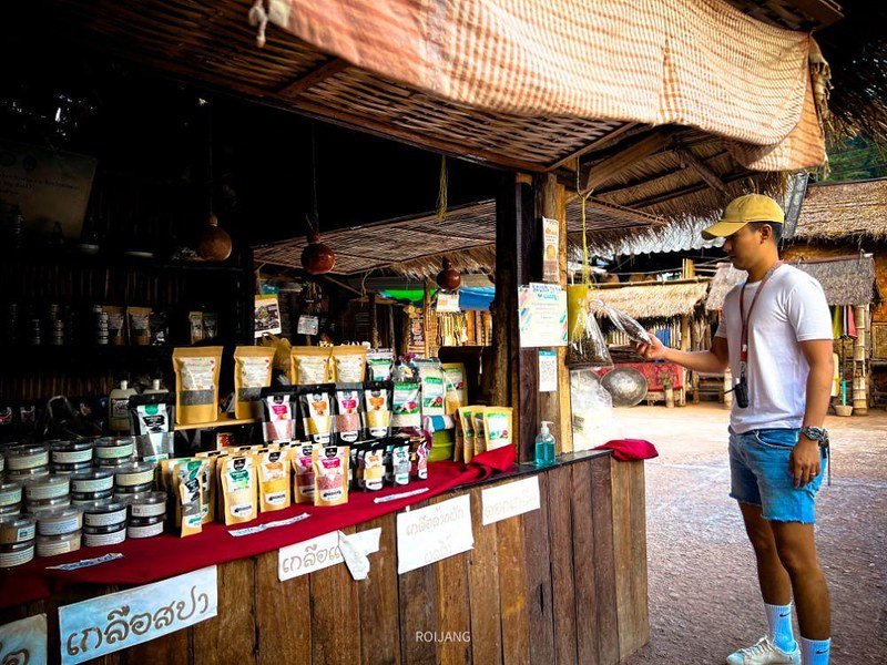 ชายคนหนึ่งสวมชุดลำลองกำลังดูสินค้าที่จัดวางบนโต๊ะที่ร้านขายของในตลาดกลางแจ้ง ซึ่งมีโครงสร้างทำจากไม้และหลังคามุงจาก สะท้อนบรรยากาศอันน่าหลงใหลที่เป็นเอกลักษณ์ของประสบการณ์การท่องเที่ยวภาคเหนือ.