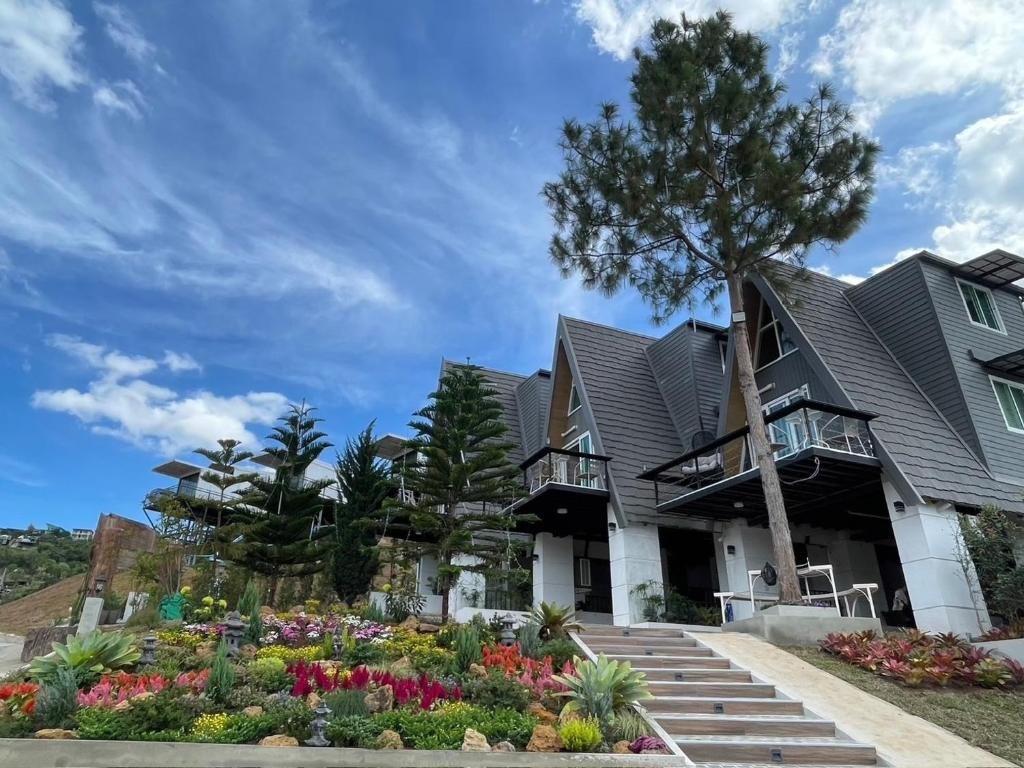 บ้านสมัยใหม่ที่มีหลังคาสามเหลี่ยม ต้นสนสูง และสวนที่จัดแต่งอย่างสวยงามด้วยดอกไม้สีสันสดใส ใต้ท้องฟ้าสีฟ้าที่มีเมฆลอยอยู่ — เหมาะอย่างยิ่งสำหรับผู้ที่กำลังหา รีสอร์ทเขาค้อ หรือมองหาที่พักเขาค้อที่สงบเงียบ ท่ามกลางความงามของธรรมชาติ.