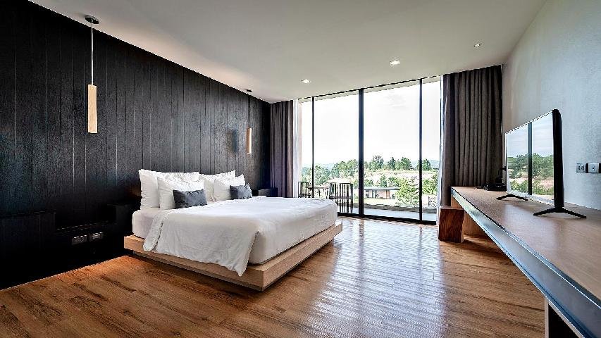 ห้องนอนสไตล์โมเดิร์นที่มีเตียงแบบแพลตฟอร์ม ชุดเครื่องนอนสีขาว พื้นไม้ ผนังสีดำเป็นจุดเด่น หน้าต่างขนาดใหญ่ โต๊ะทำงาน และทีวีจอแบนที่มองเห็นวิวพื้นที่สีเขียวด้านนอก — เหมาะอย่างยิ่งสำหรับผู้ที่กำลังมองหาที่พักเขาค้อ หรือประสบการณ์รีสอร์ทเขาค้อ ที่ผสมผสานความสบายกับวิวทิวทัศน์อันงดงาม.
