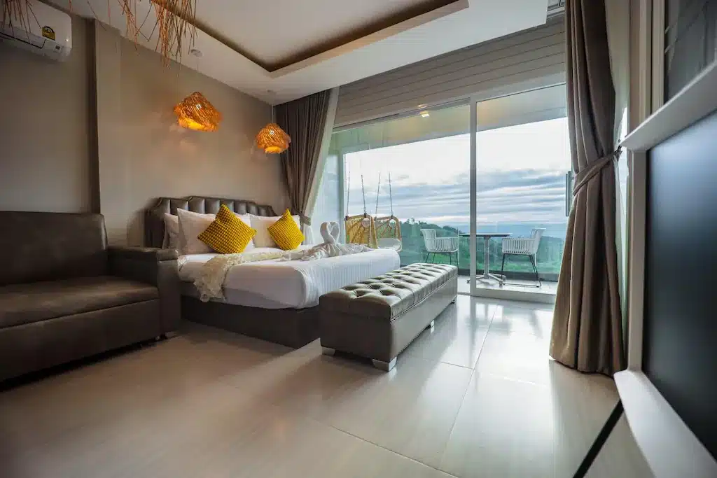 ห้องพักโรงแรมสไตล์โมเดิร์น พร้อมเตียงคู่ หมอนสีเหลือง โซฟาหนัง โต๊ะข้างเตียง และประตูแก้วขนาดใหญ่ที่เปิดออกสู่ระเบียงพร้อมพื้นที่นั่งเล่นกลางแจ้งและวิวทิวทัศน์ที่สวยงาม—เหมาะอย่างยิ่งสำหรับผู้ที่กำลังมองหาที่พักเขาค้อ หรือประสบการณ์การพักที่รีสอร์ทเขาค้อที่ไม่อาจลืมได้.