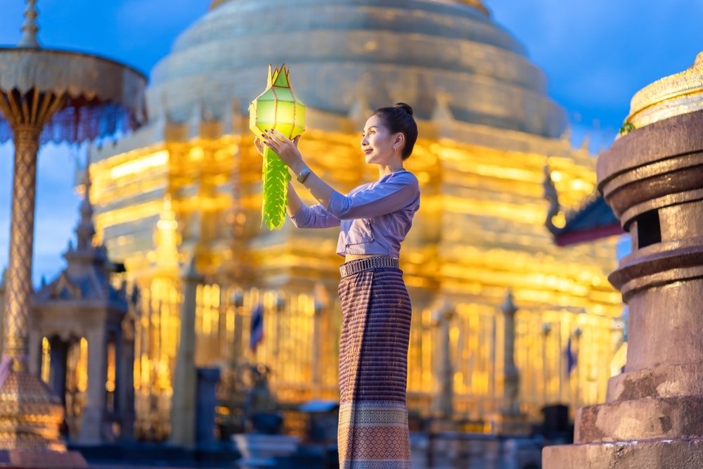 ผู้หญิงคนหนึ่งสวมชุดพื้นเมืองถือโคมไฟอยู่หน้าวัดสีทองที่สว่างไสวในยามพลบค่ำ สะท้อนบรรยากาศอันน่าหลงใหลของการท่องเที่ยวภาคเหนือและมรดกทางวัฒนธรรมอันรุ่มรวยของภูมิภาคนี้.