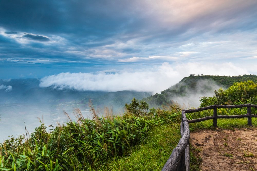 ภาพทิวทัศน์ภูเขาที่ปกคลุมด้วยหมอก พร้อมด้วยพืชพรรณเขียวชอุ่ม รั้วไม้ และท้องฟ้าที่มีเมฆครึ้มอย่างน่าทึ่งในช่วงพระอาทิตย์ขึ้นหรือพระอาทิตย์ตก — ฉากที่ชวนให้นึกถึงความงามอันสงบสุขที่พบได้ที่เที่ยวขอนแก่น ซึ่งเป็นหนึ่งในสถานที่ท่องเที่ยววเชียงคานที่สวยงามมากมาย สำหรับผู้รักธรรมชาติ.