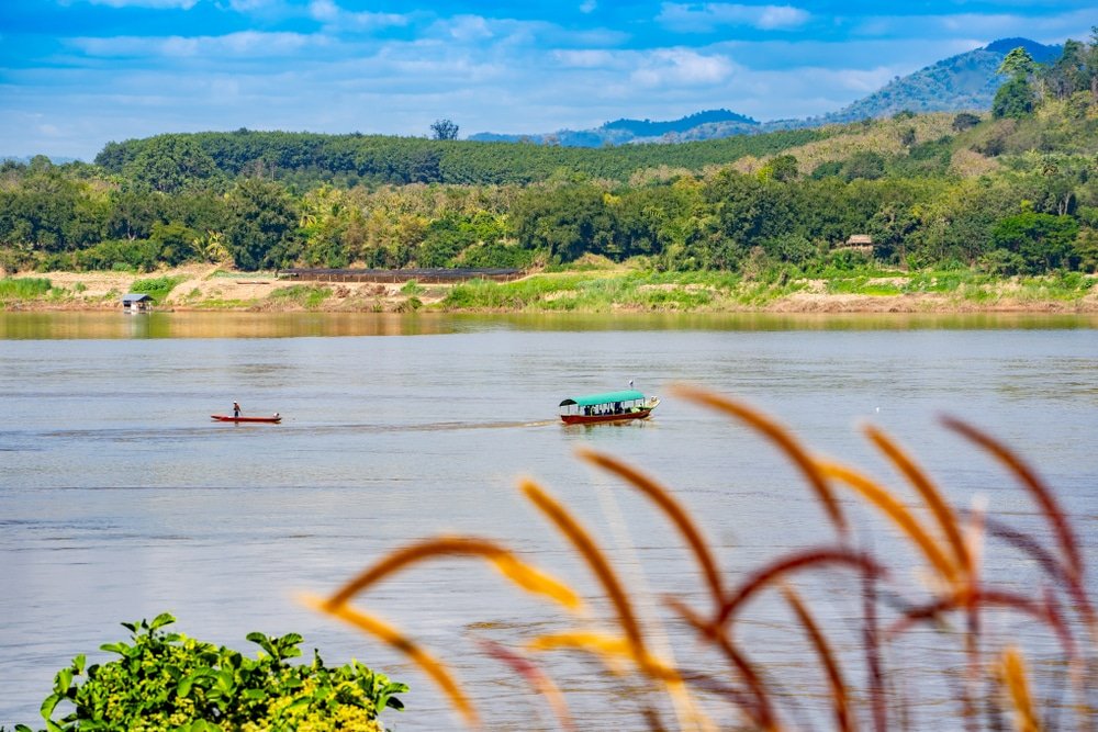 ภาพแม่น้ำที่มีเรือสองลำ หนึ่งลำใหญ่และหนึ่งลำเล็ก ล้อมรอบด้วยพืชพรรณเขียวขจีและภูเขาไกลๆ ใต้ท้องฟ้าสีฟ้า สร้างบรรยากาศความงามอันสงบของที่เที่ยวขอนแก่น ในส่วนหน้าภาพมีหญ้าสูงปรากฏให้เห็น ซึ่งเผยให้เห็นเสน่ห์ทางธรรมชาติที่มักพบได้ที่สถานที่ท่องที่เที่ยวเชียงคาน