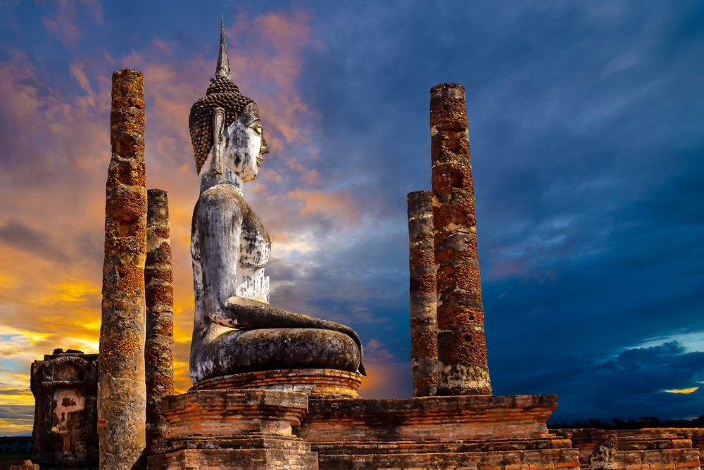 พระพุทธรูปหินขนาดใหญ่ตั้งอยู่ท่ามกลางเสาหินโบราณในยามพระอาทิตย์ตกดิน พร้อมด้วยท้องฟ้าที่งดงามตระการตาเป็นฉากหลัง — เป็นภาพอันเป็นสัญลักษณ์ที่ถ่ายทอดความงามอันไร้กาลเวลาของการท่องเที่ยวภาคเหนือ.