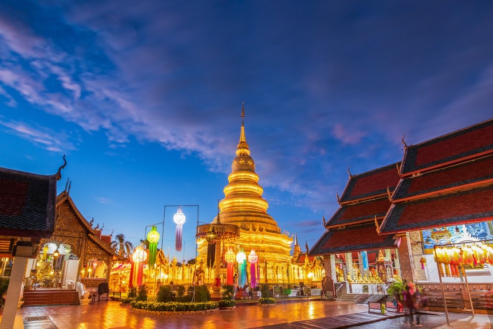 วัดสีทองพร้อมเจดีย์และโคมไฟที่ส่องแสงระยิบระยับยามพลบค่ำ ล้อมรอบด้วยอาคารแบบดั้งเดิมใต้ท้องฟ้าสีฟ้าและม่วง—ภาพอันน่าหลงใหลที่สะท้อนแก่นแท้ของการท่องเที่ยวภาคเหนือ สำหรับผู้ที่แสวงหาประสบการณ์ทางวัฒนธรรมที่ไม่อาจลืมเลือนในภาคเหนือของประเทศไทย.