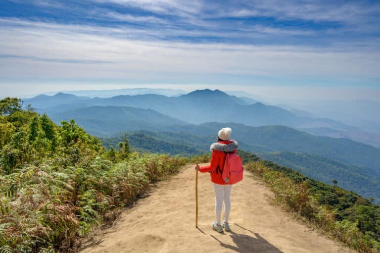 มีชายคนหนึ่งสวมแจ็กเก็ตสีแดงและหมวกสีขาว ยืนอยู่บนทางดิน ถือไม้เท้า มองออกไปยังภูเขาสีเขียวใต้ท้องฟ้าสีฟ้าที่มีเมฆกระจายตัวอยู่บ้าง—ภาพนี้ถ่ายทอดความงามอันสงบของเที่ยวภาคเหนือได้อย่างสมบูรณ์แบบ.