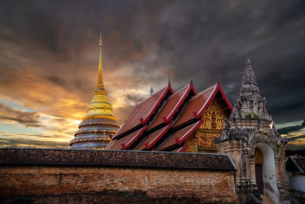 เจดีย์สีทองและอาคารวัดที่ตกแต่งอย่างวิจิตรบรรจงในยามพระอาทิตย์ตกดิน พร้อมด้วยเมฆที่ดูน่าทึ่งบนท้องฟ้า สร้างบรรยากาศที่น่าหลงใหลของการท่องเที่ยวภาคเหนือ ซึ่งน่าจะเป็นในประเทศไทย.