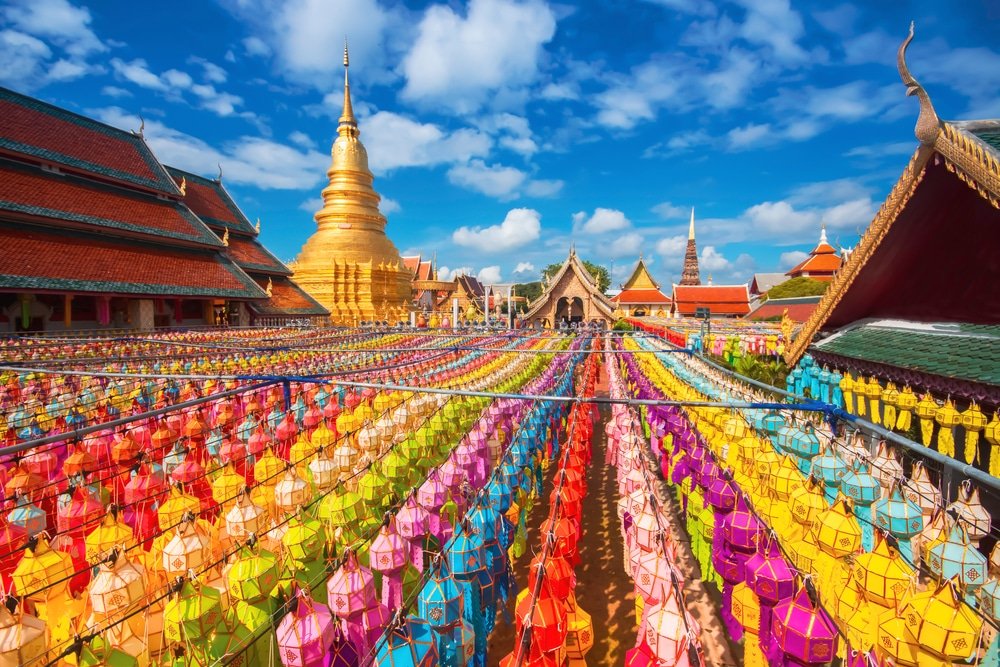 โคมกระดาษสีสันสดใสถูกจัดเรียงเป็นแถวอยู่ด้านหน้าเจดีย์สีทองและอาคารวัดต่าง ๆ ใต้ท้องฟ้าสีฟ้าสดใสที่มีเมฆลอยอยู่ สร้างบรรยากาศที่คึกคักและเต็มไปด้วยความรื่นเริง ซึ่งถ่ายทอดเสน่ห์ของการท่องเที่ยวภาคเหนือได้อย่างสมบูรณ์แบบ.