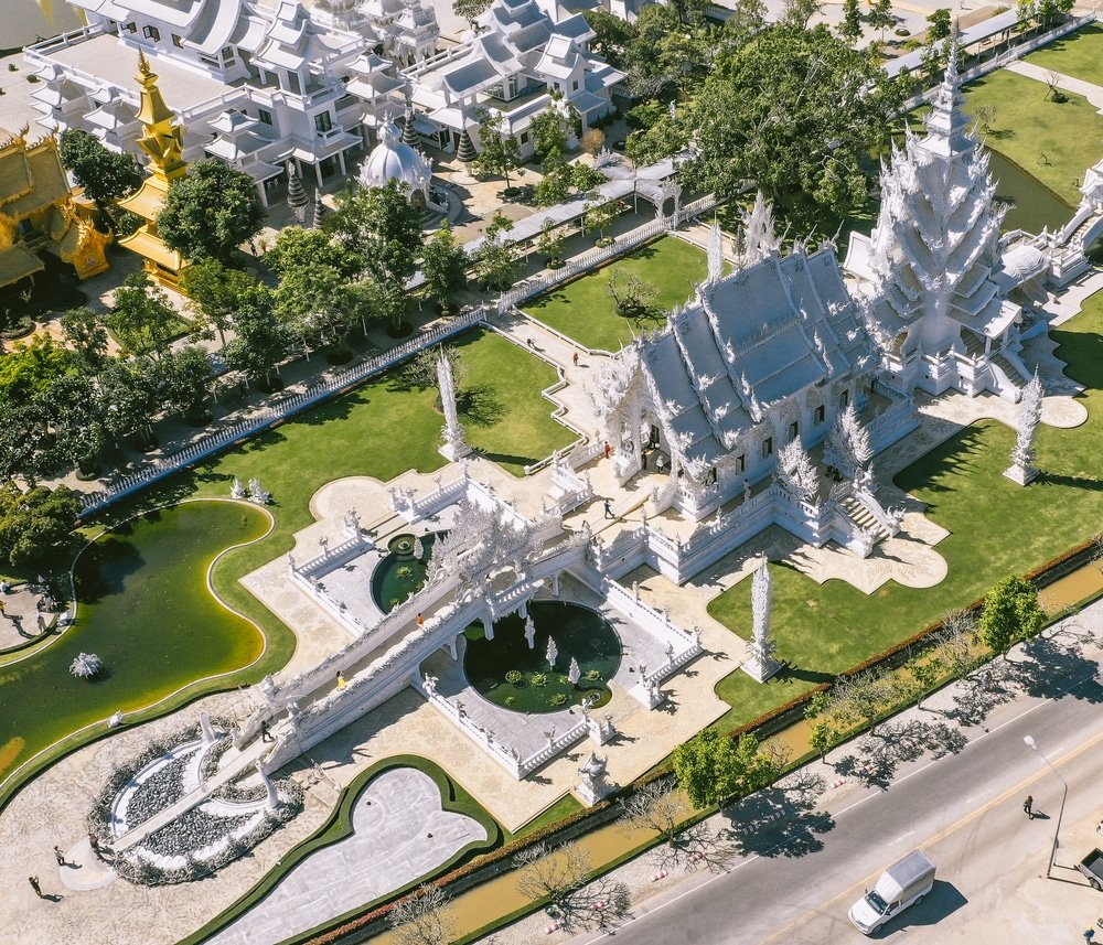 ภาพจากมุมสูงของวัดขาว (วัดร่องขุน) ในจังหวัดเชียงราย ประเทศไทย ซึ่งแสดงให้เห็นโครงสร้างสีขาวที่ประดับตกแต่งอย่างวิจิตร สนามหญ้าสีเขียว สระน้ำ และทางเดินรอบๆ — เป็นจุดท่องเที่ยวที่ไม่ควรพลาดสำหรับใครก็ตามที่ต้องการท่องเที่ยวภาคเหนือและสัมผัสความงามอันเป็นเอกลักษณ์ของภาคเหนือประเทศไทย.
