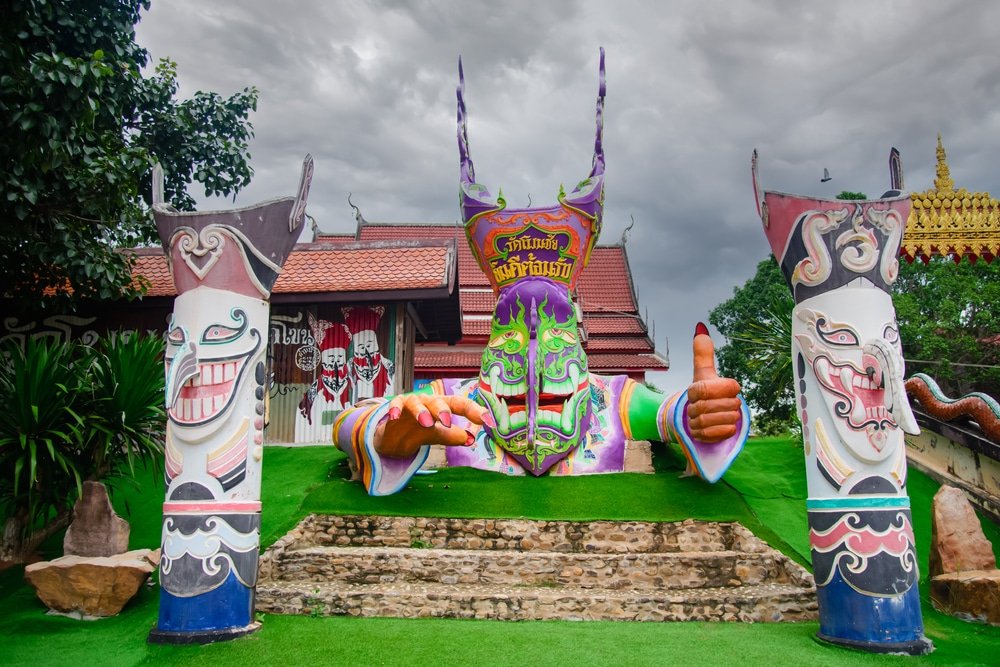 รูปปั้นหน้ากากขนาดใหญ่และสีสันสดใส พร้อมด้วยมือที่ยื่นออกมาและเสาที่ตกแต่งอย่างวิจิตร ตั้งอยู่บนสนามหญ้าสีเขียวด้านหน้าอาคารแบบไทยดั้งเดิม ใต้ท้องฟ้าที่มีเมฆครึ้ม ทำให้ที่นี่เป็นสถานที่ที่ไม่ควรพลาดสำหรับใครก็ตามที่กำลังมองหาสถานที่ท่องเที่ยวในขอนแก่น หรือสถานที่ท่องเที่ยวที่ไม่เหมือนใครในเที่ยวเชียงคาน 