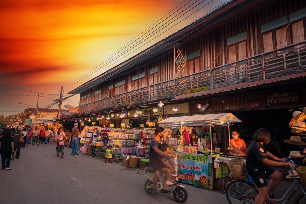 ตลาดถนนที่คึกคักเต็มไปด้วยแผงขายสินค้าและผู้ซื้อเริ่มคึกคักขึ้นหน้าอาคารไม้เมื่อพระอาทิตย์ตกดิน — เป็นภาพที่สวยงามและเป็นเอกลักษณ์ของสถานที่ท่องเที่ยวขอนแก่น บางคนขี่จักรยาน ส่วนคนอื่นเดินผ่านร้านค้าต่าง ๆ พร้อมซึมซับบรรยากาศที่คึกคัก ซึ่งทำให้ที่เที่ยวเชียงคาน