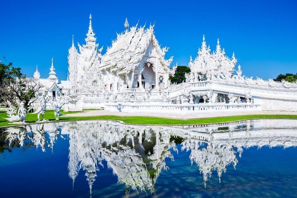 วัดสีขาวที่ตกแต่งอย่างวิจิตรด้วยลวดลายแกะสลักที่ละเอียดอ่อนและยอดแหลมที่สะท้อนภาพในสระน้ำใต้ท้องฟ้าสีฟ้าใส ล้อมรอบด้วยหญ้าสีเขียวและต้นไม้—เหมาะอย่างยิ่งสำหรับผู้ที่ต้องการท่องเที่ยวภาคเหนือและค้นพบความงามอันเป็นเอกลักษณ์ของสถานที่ท่องเที่ยวภาคเหนือ.