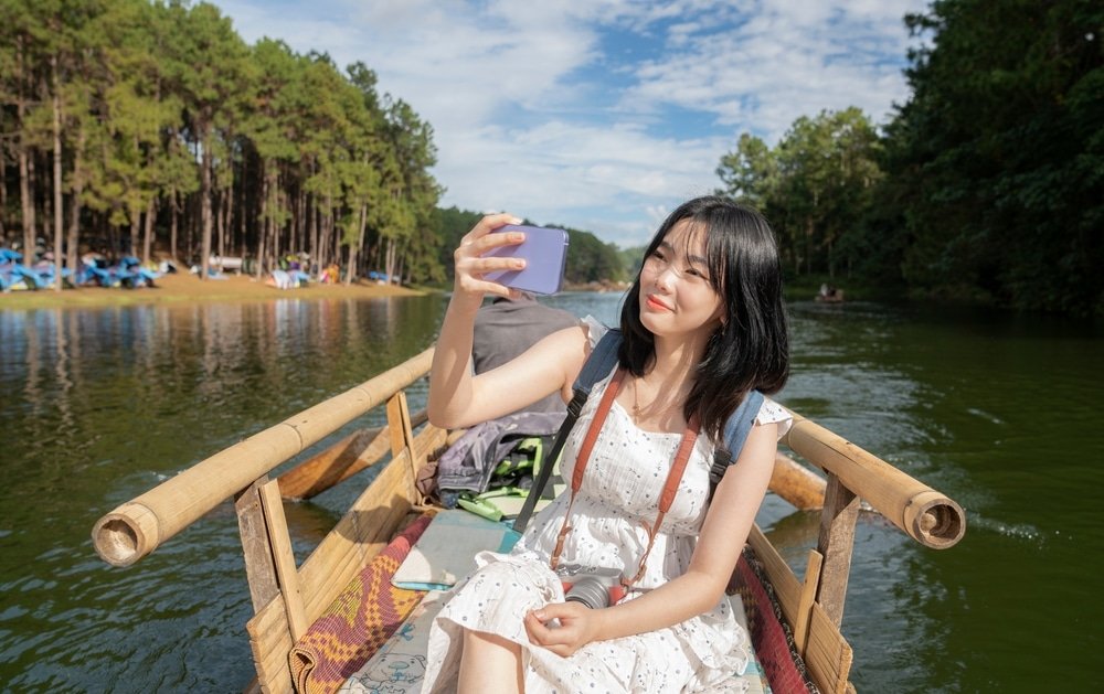 ผู้หญิงคนหนึ่งสวมชุดสีขาวนั่งอยู่ในเรือไม้บนแม่น้ำ กำลังถ่ายเซลฟี่ด้วยโทรศัพท์มือถือ ต้นสนและพื้นที่ตั้งแคมป์ปรากฏให้เห็นตามริมแม่น้ำใต้ท้องฟ้าสีฟ้า ซึ่งถ่ายทอดความงามอันสงบของที่เที่ยวภาคเหนือ สำหรับผู้ที่ชื่นชอบการสำรวจภาคเหนือของประเทศไทย.