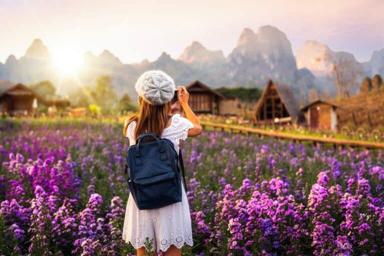 มีคนหนึ่งสะพายเป้และสวมชุดสีขาว ยืนอยู่ท่ามกลางทุ่งดอกไม้สีม่วง กำลังถ่ายภาพทิวทัศน์ภูเขาและบ้านไม้สไตล์ชนบทในยามพระอาทิตย์ขึ้น — เป็นช่วงเวลาที่สมบูรณ์แบบสำหรับใครก็ตามที่ต้องการเที่ยวภาคเหนือ และดื่มด่ำกับความงามตามธรรมชาติและเสน่ห์ของภาคเหนือของไทย.