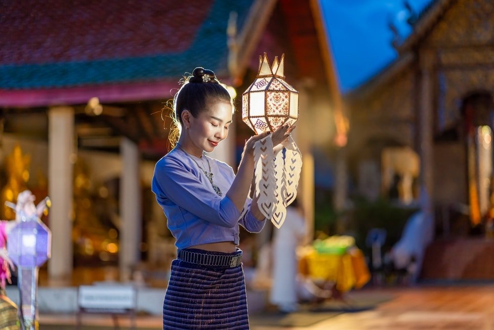 ผู้หญิงคนหนึ่งสวมชุดพื้นเมืองถือโคมประดับอยู่กลางแจ้งในช่วงพลบค่ำ โดยมีโครงสร้างวัดปรากฏอยู่เบื้องหลัง — ภาพที่ถ่ายทอดบรรยากาศอันน่าหลงใหลของการท่องเที่ยวภาคเหนือได้อย่างงดงาม.