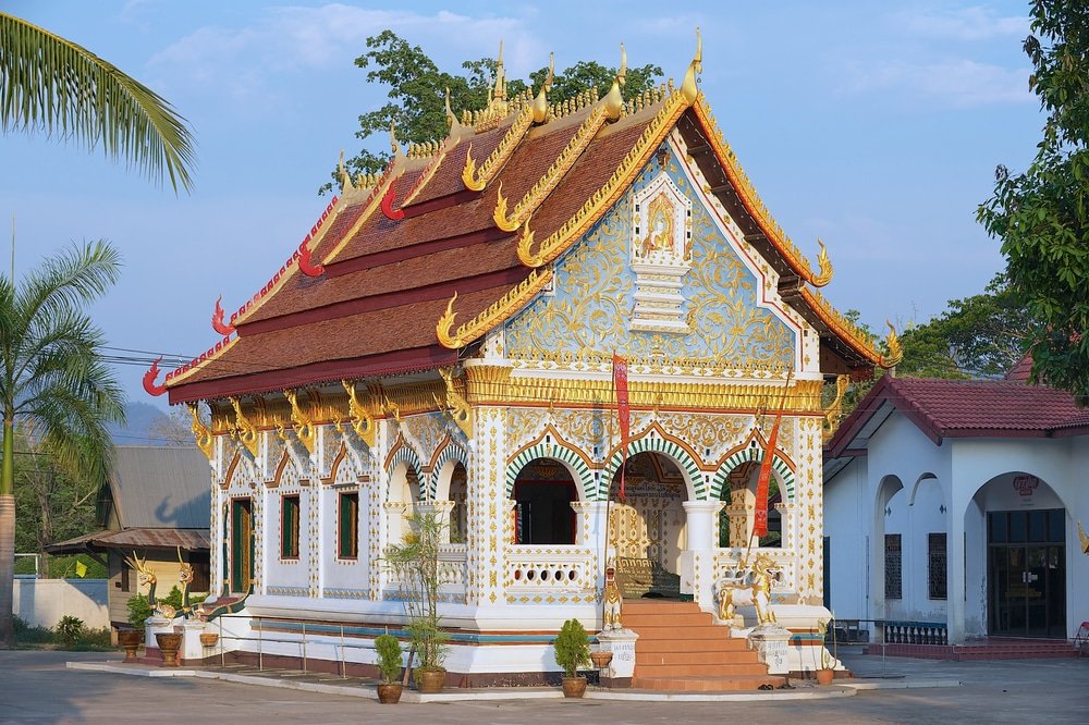 วัดพุทธขนาดเล็กที่ตกแต่งอย่างวิจิตรด้วยรายละเอียดประดับสีทอง หลังคาสีแดง และผนังสีขาว ล้อมรอบด้วยต้นไม้ในกระถางและอาคารใกล้เคียง ภายใต้ท้องฟ้าแจ่มใส — สถานที่อันสงบแห่งนี้คือหนึ่งในที่เที่ยวเชียงคานมีเสน่ห์ เหมาะอย่างยิ่งสำหรับนักท่องเที่ยวที่ต้องการสัมผัสวัฒนธรรมท้องถิ่น.