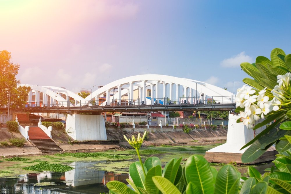 สะพานโค้งสีขาวทอดข้ามแม่น้ำ โดยมีพืชพรรณและดอกไม้สีเขียวอยู่เบื้องหน้า ภายใต้ท้องฟ้าที่มีเมฆบางส่วน ทำให้ที่นี่กลายเป็นหนึ่งในสถานที่ท่องเที่ยวภาคเหนือที่น่าหลงใหล สำหรับผู้ที่ต้องการสำรวจความงามของธรรมชาติและสถานที่สำคัญอันเป็นเอกลักษณ์ของภาคเหนือของประเทศไทย.