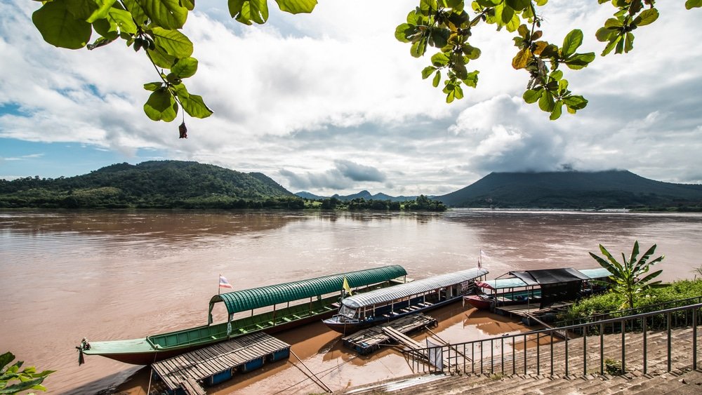 เรือยาวสามลำจอดอยู่ริมฝั่งแม่น้ำที่เต็มไปด้วยโคลน โดยมีภูเขาสีเขียวและท้องฟ้าครึ้มเป็นฉากหลัง ใบไม้ล้อมรอบส่วนบนของภาพ ช่วยถ่ายทอด ที่เที่ยวเชียงคานบรรยากาศอันสงบของขอนแก่นที่เที่ยว สำหรับผู้ที่ต้องการค้นพบสถานที่ใหม่ๆ ในภูมิภาคที่มีเสน่ห์แห่งนี้.