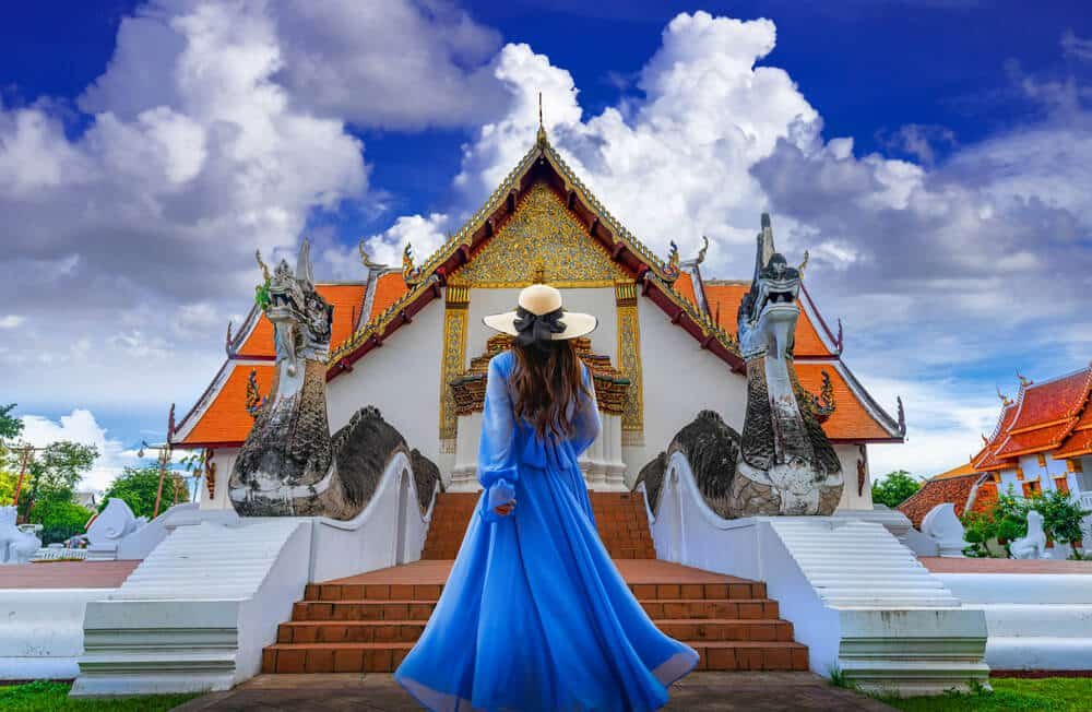 ผู้หญิงคนหนึ่งสวมชุดสีฟ้าและหมวก ยืนหันหน้าสู่วัดไทยแบบดั้งเดิมที่มีลวดลายประดับตกแต่งอย่างวิจิตร พร้อมด้วยรูปปั้นนาคสองตัวตั้งอยู่ข้างบันได ใต้ท้องฟ้าที่สดใส — ภาพที่ถ่ายทอดความเสน่ห์ของการท่องเที่ยวภาคเหนือ.