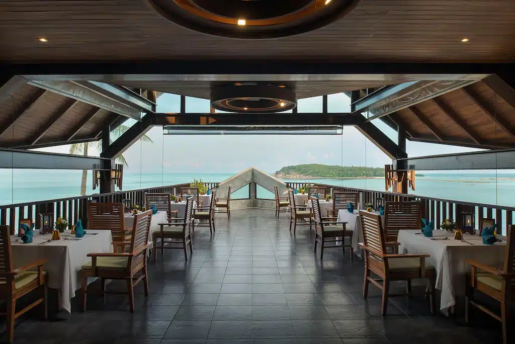 ระเบียงร้านอาหารกลางแจ้งที่ที่พักเกาะสมุย ซึ่งมีโต๊ะจัดไว้สำหรับมื้อเย็น พร้อมวิวทะเลและเกาะที่อยู่ไกลๆ.