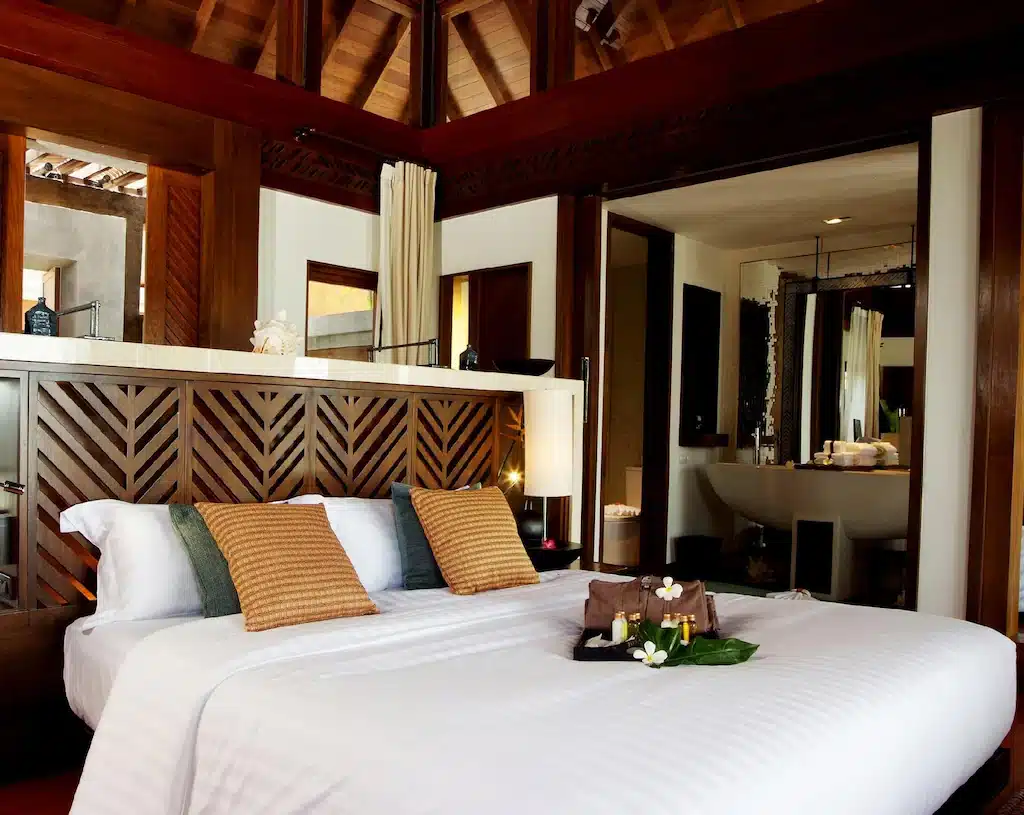 ห้องพักในโรงแรมแห่งนี้ ที่พักสมุย มีหัวเตียงทำจากไม้ เตียงสีขาวพร้อมหมอนตกแต่งสีน้ำตาล และห้องน้ำแบบเปิดที่มีอ่างล้างหน้าทำจากหิน.