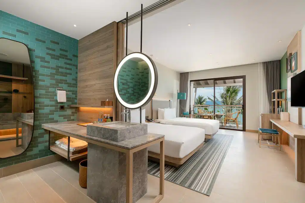 ห้องพักในโรงแรมแห่งนี้ ที่พักเกาะสมุย มีโต๊ะแต่งหน้าทำจากหินพร้อมกระจกที่มีไฟส่องสว่าง เตียงสองเตียง และประตูแก้วที่เปิดออกสู่ระเบียง ซึ่งสามารถมองเห็นวิวต้นปาล์มและทะเล.