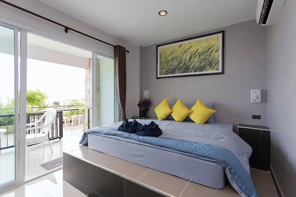 ห้องพักในโรงแรมที่มีเตียงพร้อมหมอนสีเหลืองสามใบ ผ้าขนหนูสีเข้มสองผืน และภาพวาดทุ่งข้าวสาลีบนผนัง พร้อมประตูเลื่อนที่นำไปสู่ระเบียง ที่พักราคาประหยัดบนเกาะลันแห่งนี้.