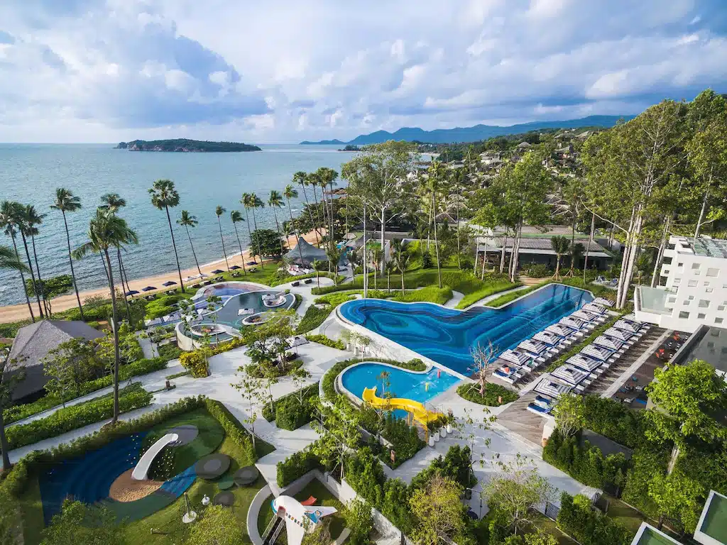 ภาพจากอากาศของรีสอร์ทริมชายหาดบนเกาะสมุย ซึ่งมีสระว่ายน้ำหลายแห่ง สนามเล่นเด็ก และพื้นที่สีเขียวที่ร่มรื่นอยู่ข้างชายหาด.
