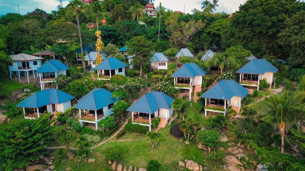 ภาพจากด้านบนของรีสอร์ทที่มีบังกะโลสีขาวและหลังคาสีฟ้า ตั้งอยู่บนเนินเขาที่ร่มรื่นและเต็มไปด้วยต้นไม้ ที่พักเกาะสมุยแห่งนี้.
