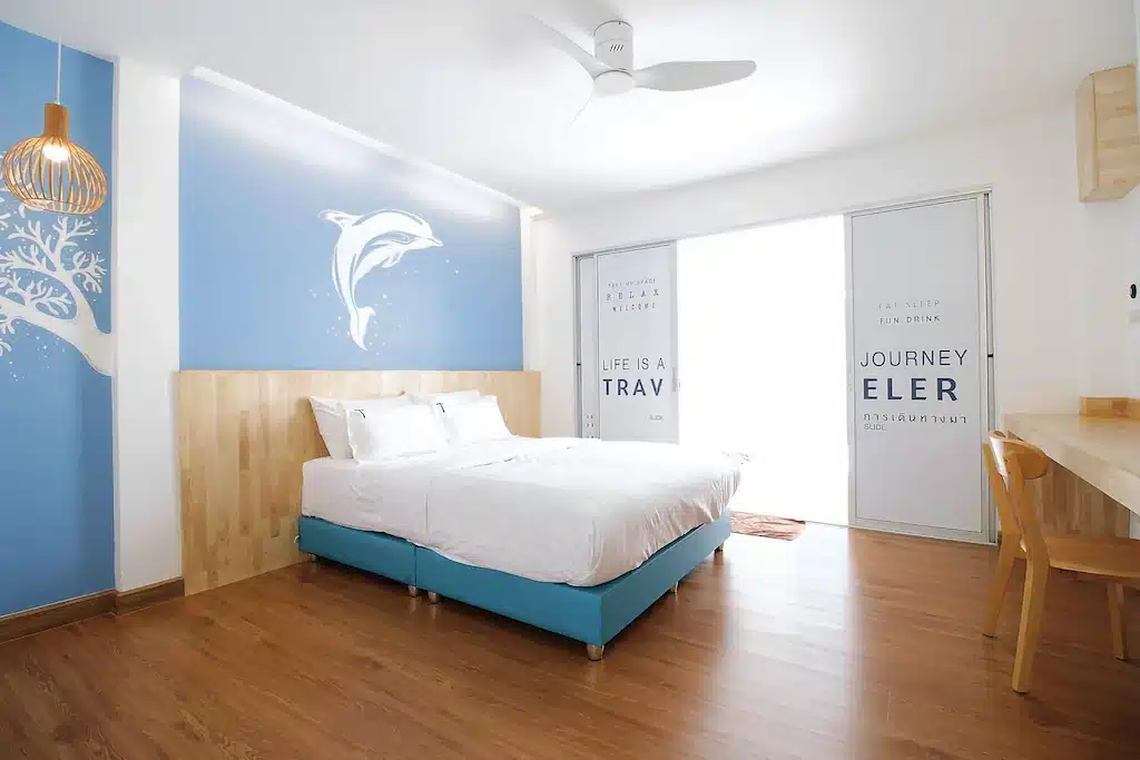 ห้องพักในโรงแรมที่มีเตียงควีนตั้งอยู่หน้าผนังที่มีภาพจิตรกรรมฝาผนังรูปปลาโลมา พื้นไม้ และประตูแก้ว; ที่พักเกาะล้านติดทะเลที่สะดวกสบาย.
