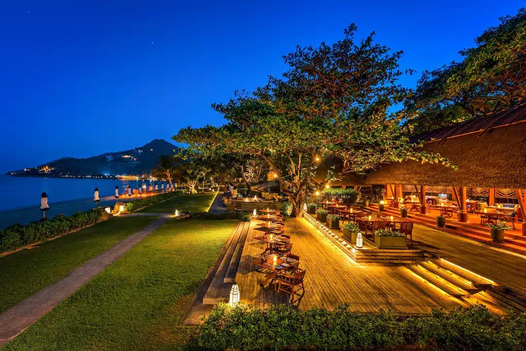 ที่นั่งรับประทานอาหารกลางแจ้งบนดาดฟ้าไม้ของรีสอร์ทบนเกาะสมุย ตั้งอยู่ข้างสนามหญ้าและชายหาดในช่วงพลบค่ำ.