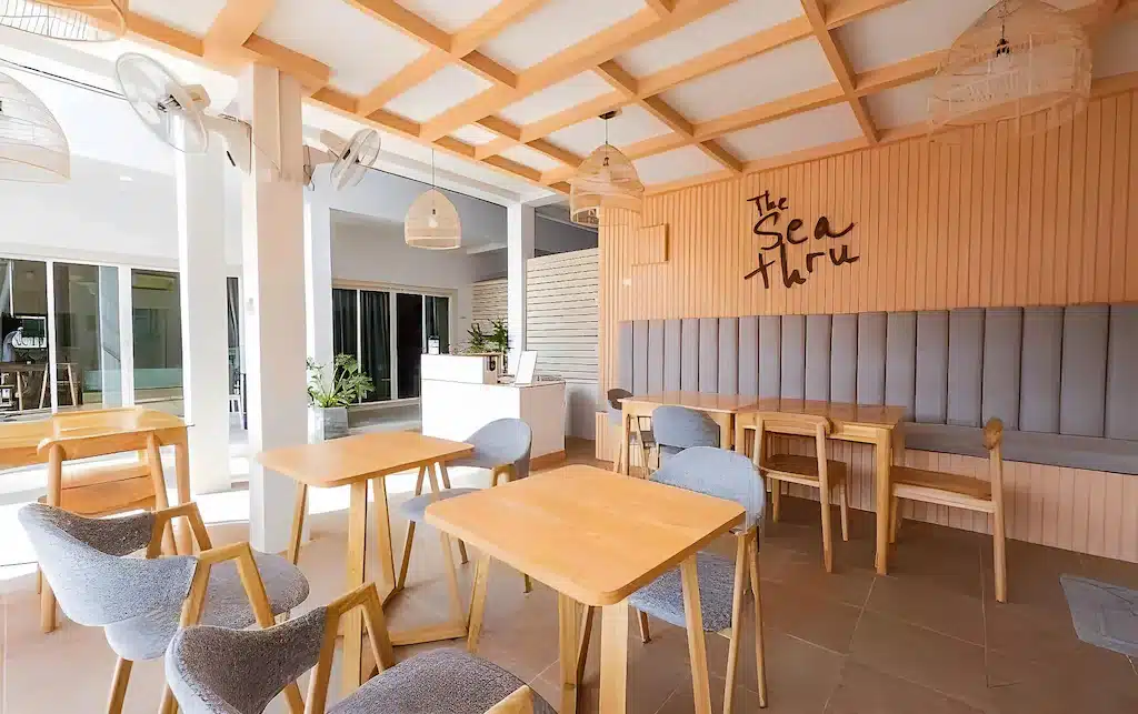 ห้องรับประทานอาหารที่มีโต๊ะไม้ เก้าอี้หุ้มผ้าสีเทา และเก้าอี้ยาวติดผนังที่มีป้าย “The Sea Thru.” เป็นสถานที่ที่สมบูรณ์แบบสำหรับผู้ที่กำลังมองหาที่พักเกาะล้าน.