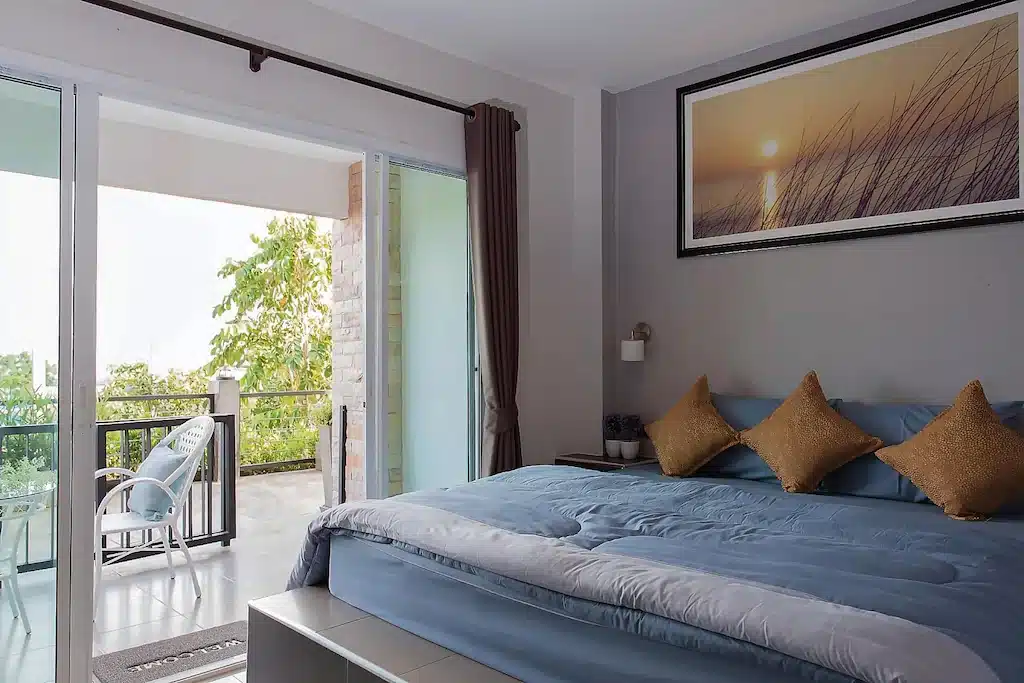 ห้องพักในโรงแรมที่มีเตียงคลุมผ้าสีฟ้า หมอนสีทอง และระเบียงที่มีพื้นที่นั่งเล่นกลางแจ้ง ซึ่งเหมาะสำหรับผู้ที่กำลังมองหาที่พักราคาประหยัดบนเกาะลัน.