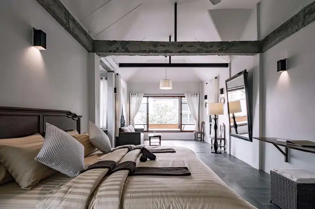 ห้องนอนในโรงแรมที่มีเตียงขนาดใหญ่ กระจกติดผนัง และพื้นที่นั่งเล่นข้างหน้าต่างที่มองเห็นต้นไม้ เหมาะอย่างยิ่งสำหรับผู้ที่กำลังวางแผนเดินทางไปเที่ยวเชียงราย.