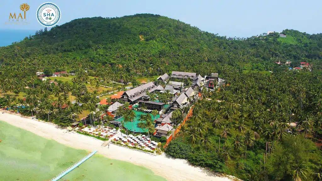 ภาพจากมุมสูงของ Mai Samui Beach Resort ซึ่งเป็นที่พักชั้นนำบนเกาะสมุย ที่มีสระว่ายน้ำหลายแห่งและอาคารต่าง ๆ ตั้งอยู่ระหว่างเนินเขาเขียวชอุ่มและชายหาดทรายขาว.