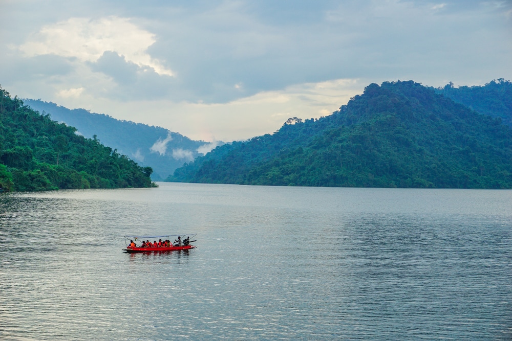 เรือเล็กสีแดงที่บรรทุกผู้โดยสารกำลังแล่นข้ามแม่น้ำกว้าง ซึ่งล้อมรอบด้วยภูเขาที่ปกคลุมด้วยป่า ในจังหวัดนครนายก ซึ่งเป็นสถานที่ท่องเที่ยวยอดนิยม.