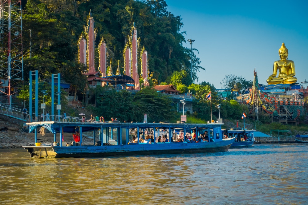 เรือโดยสารสีฟ้าตัวยาวแล่นไปตามแม่น้ำผ่านเนินเขาที่มีพระพุทธรูปสีทองตั้งอยู่ ซึ่งเป็นจุดเด่นสำหรับผู้ที่มาเที่ยวที่เที่ยวเชียงราย.