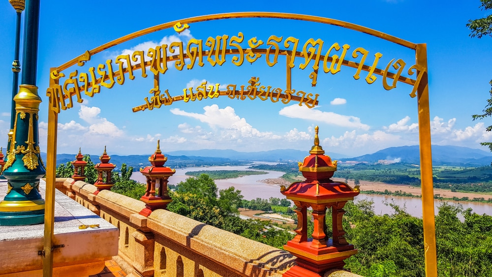 ประตูโค้งทำจากโลหะสีทองที่มีอักษรไทยตั้งอยู่บนระเบียงที่มองเห็นแม่น้ำไหลคดเคี้ยวผ่านภูเขาใต้ท้องฟ้าสีคราม ซึ่งเป็นจุดเด่นสำหรับผู้ที่มาเที่ยวเชียงราย.