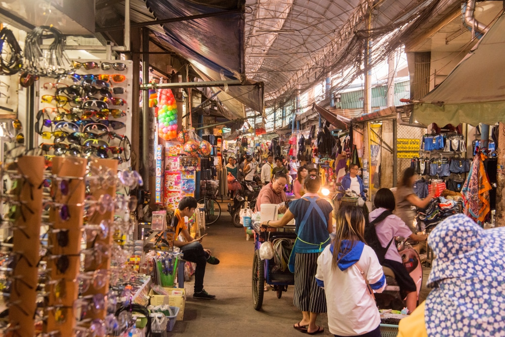 ผู้ซื้อเดินผ่านตลาดริมถนนที่คับแคบและแออัดในเชียงราย ซึ่งมีแผงขายแว่นกันแดดและเสื้อผ้าเรียงรายอยู่เต็มไปหมด.