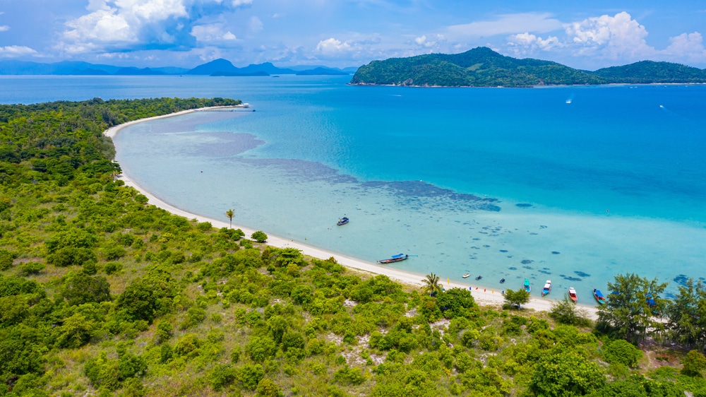 ภาพจากมุมสูงของชายหาดทรายโค้งที่ติดกับป่าเขียวชอุ่ม ซึ่งทำให้นึกถึงสถานที่ท่องเที่ยวที่สวยงามใน ที่เที่ยวเกาะสมุย 2026  พร้อมด้วยเรือที่จอดอยู่ในน้ำสีเทอร์ควอยซ์ใกล้เกาะขนาดใหญ่.